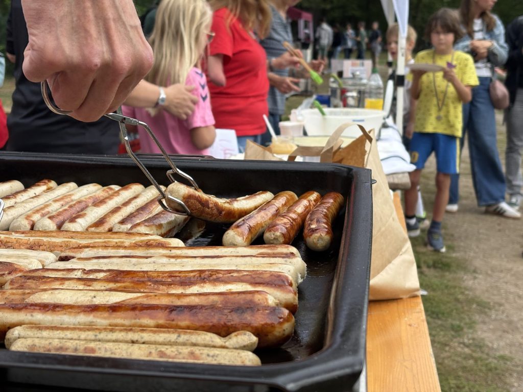 Bratwürste liegen auf dem Grill, im Hintergrund warten Kinder